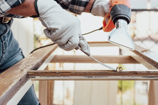 Adult carpenter wearing protective leather gloves, using a heat gun and a wood scraper to remove paint from an old wooden window. Construction industry, carpentry. Restoration. 