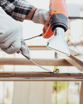 Adult carpenter wearing protective leather gloves, using a heat gun and a wood scraper to remove paint from an old wooden window. Construction industry, carpentry. Restoration. 