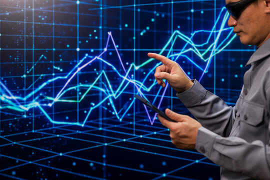 Technician wearing safety glasses uses a tablet and points at glowing financial charts on a digital grid screen, showing data analysis and market trends.