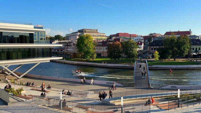 Svisloch River embankment in Minsk, with pedestrian Lavsky Bridge and Trinity Suburb. Pleasure boat speeds through water while buildings background. Belarus, Minsk, September 16, 2026