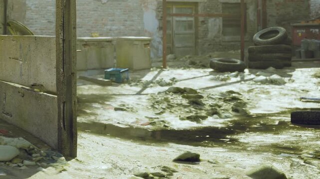 A neglected alley lies in disrepair, with muddy patches and scattered tires. Sunlight filters through the decay, illuminating the forgotten space and its surrounding debris.