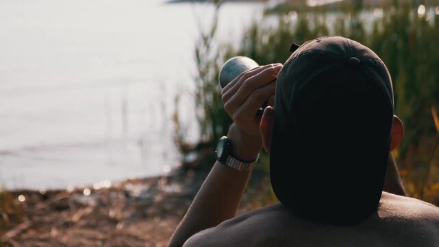 Shirtless person in a cap adjusts binoculars near grassy shoreline during late afternoon light. Precision optical equipment enhances wildlife observation against warm backlighting.