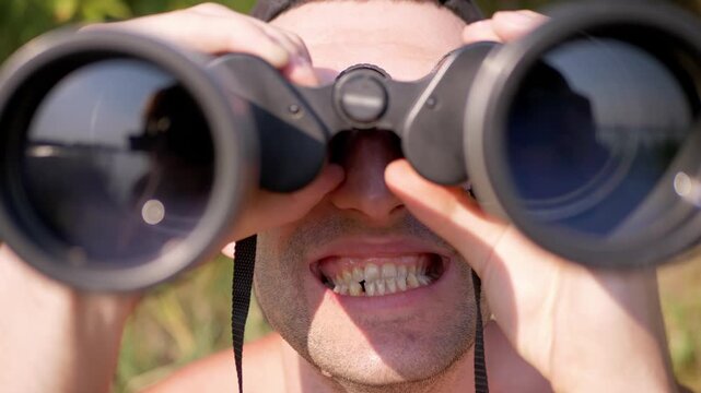 A young man holding black binoculars looks ahead with a wide, toothy, quirky grimace outdoors. Bright natural sunlight illuminates his expressive face and irregular dental smile.