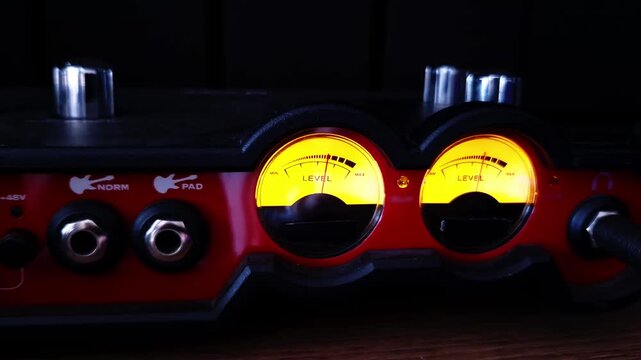 Illuminated analog VU meters glow bright amber on a red audio interface panel in a dim recording studio. Metallic dials and input jacks highlight professional sound engineering hardware.