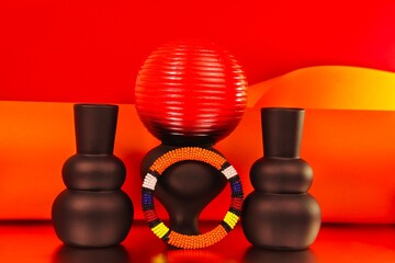 ibrant African beaded bracelet featuring bold orange, yellow, red, blue and white patterns, styled with sculptural vases against a striking red and orange backdrop for a colourful fashion still life. © Valerie
