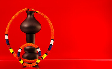ibrant African beaded bracelet featuring bold orange, yellow, red, blue and white patterns, styled with sculptural vases against a striking red and orange backdrop for a colourful fashion still life. © Valerie