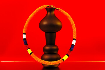 ibrant African beaded bracelet featuring bold orange, yellow, red, blue and white patterns, styled with sculptural vases against a striking red and orange backdrop for a colourful fashion still life. © Valerie