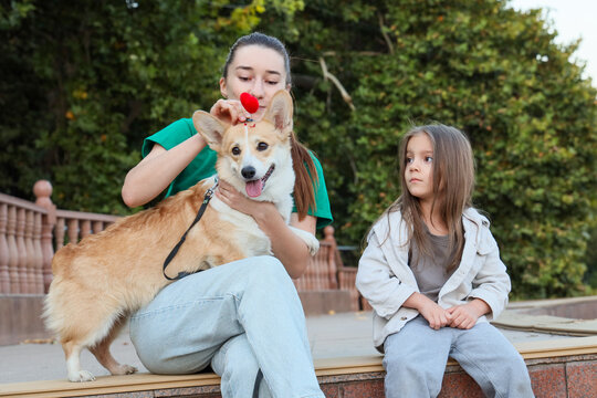 Young Woman Holding Corgi Dog With Heart Toy Beside Curious Little Girl On Outdoor Steps In Park, Showing Playful Family Bond, Pet Care, And Warm Summer Interaction Against Soft Green Background