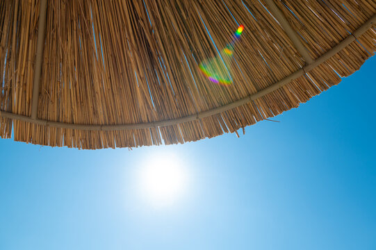 Sunlight shines through a blue sky beneath the edge of a thatched beach umbrella