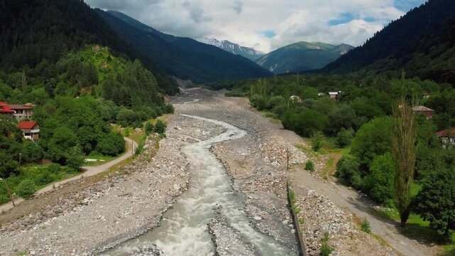 The aerodrone flew over a fast-flowing mountain river and stream in a rocky valley surrounded by green forests and snow-capped peaks of the Caucasus Mountains in Racha, Georgia