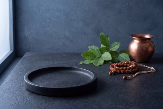 Maha Shivaratri product background with bilva leaves, rudraksha beads and a copper vessel beside an empty dark stone display