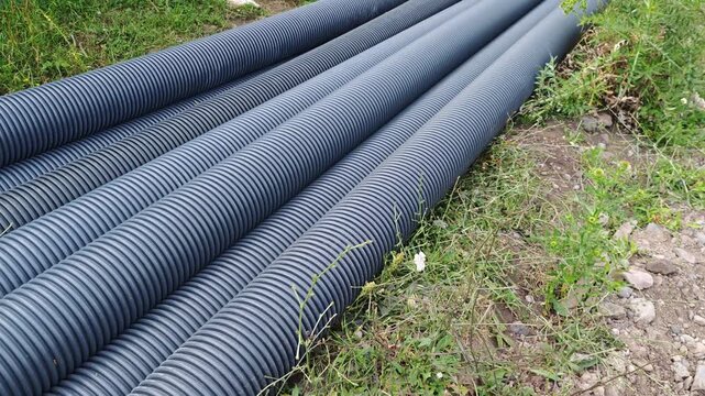 Large diameter black corrugated plastic pipes lying on the grass and ground at a construction site, prepared for installation of drainage, water supply, or sewerage systems