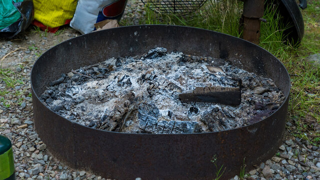 Rusty metal fire ring filled with gray ash and charred logs outdoors in a grassy gravel setting