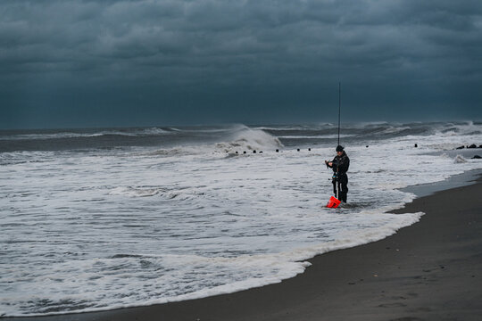 An angler fishes beside a curving surf line on the New York coast. A person holds a rod beside a red bucket as white foam spreads across the beach under gray clouds.