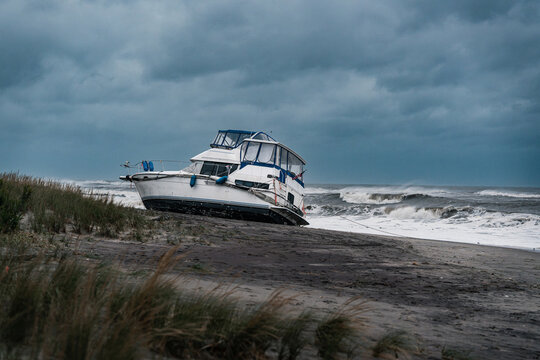 A beached boat sits beside breaking waves on the New York coast. A white cabin cruiser sits left of center on dark sand with grass below and clouds above.