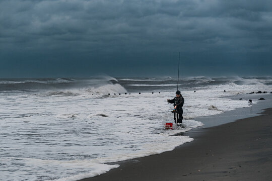 An angler faces a band of breaking waves on the New York coast. A person holds a rod on the wet shoreline with a red bucket at their feet and clouds overhead.
