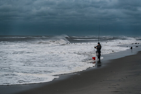 A fishing rod rises above the surf on the New York coast. An angler stands right of center beside a red bucket as white foam reaches the dark sand.