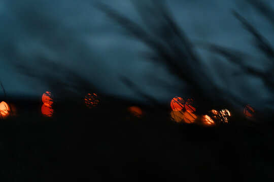 Coastal grass blurs against amber lights beneath a teal sky. Diagonal silhouettes cross glowing circles in an out of focus shoreline scene at dusk.