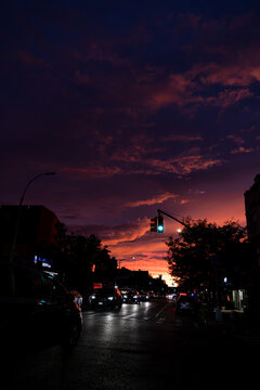 Traffic fills a New York street beneath a purple evening sky. Vehicles and a green signal occupy the lower frame beneath red clouds and an orange horizon.
