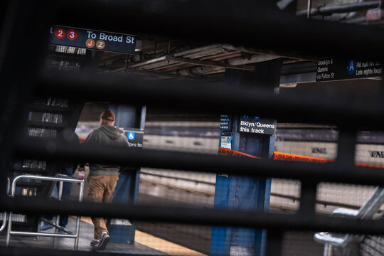A New York subway platform appears through dark railings. A passenger stands near a route sign behind horizontal bars with blue columns and tiled walls.