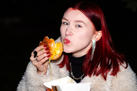 Happy girl eating take away hamburger outside. Close up of gen Z girl eating junk food while feeling excited. Harsh flash spontaneous photo. Retro inspired urban lifestyle