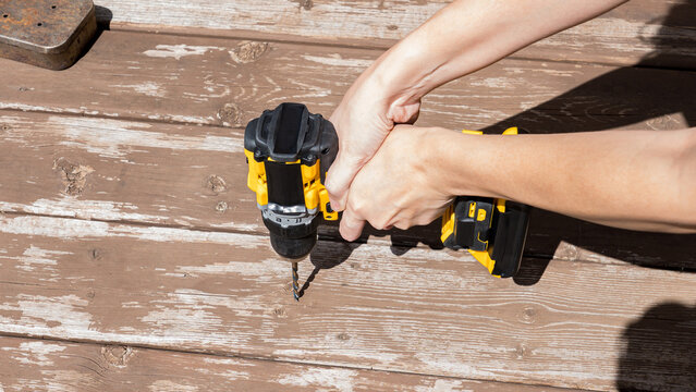 Hands holding a cordless drill above weathered wooden planks with scattered tools in a bright outdoor repair workspace