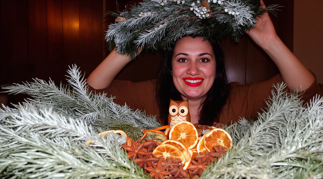    Candid portrait of a woman playfully holding a large winter wreath with dried oranges and a wooden owl. Dark wood background. Joyful holiday spirit.
