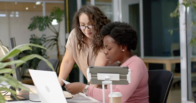 Diverse female coworkers leaning in pointing at papers and white laptop urging seated woman to type