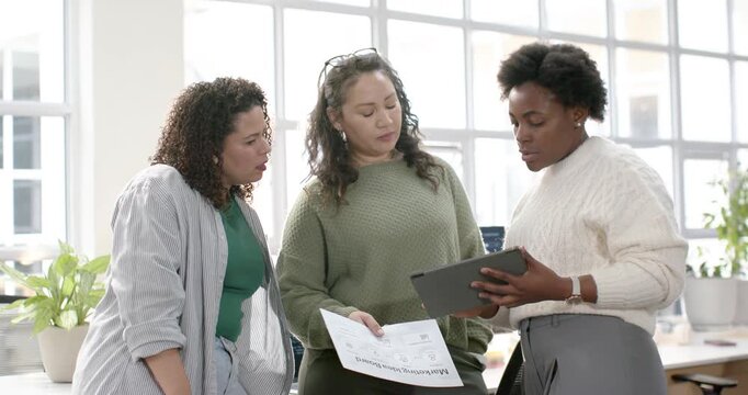 Diverse women team in sunlit office, right woman tapping tablet showing printout for slide review