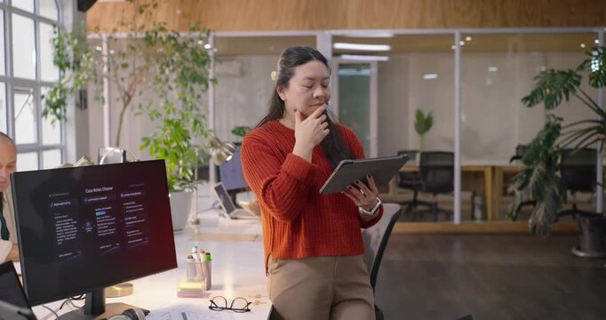 Chinese woman wearing rust top at desk in open plan office, tapping tablet prompted by dashboard