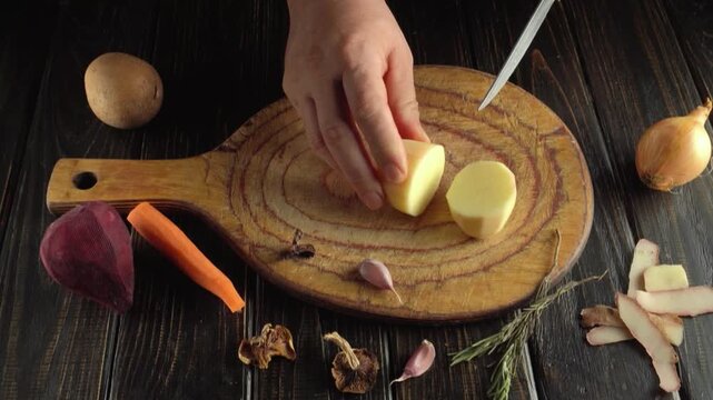 Hands slicing a peeled potato on a rustic wooden cutting board surrounded by vegetables, garlic, herbs, and kitchen ingredients in a top view food preparation scene