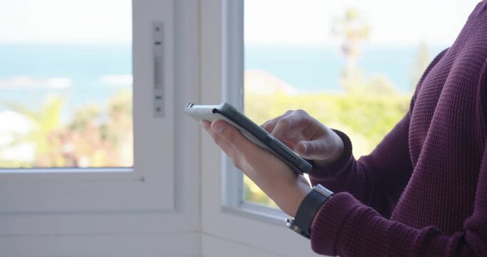 Hands are holding smartphone and are tapping, swiping and scrolling to check messages near window