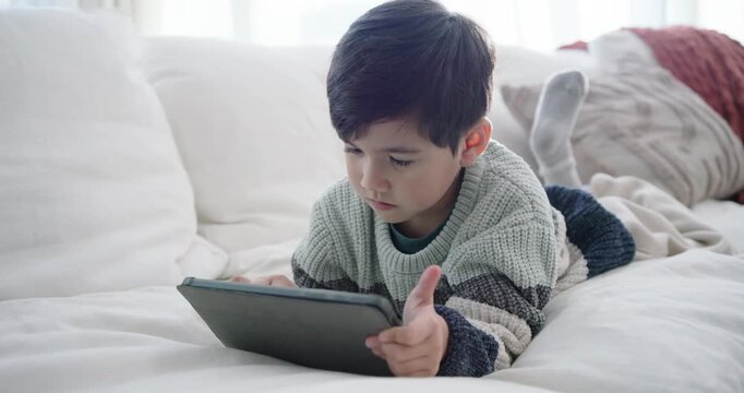 Asian male child lying stomach tapping tablet, following prompts on big sofa in striped sweater