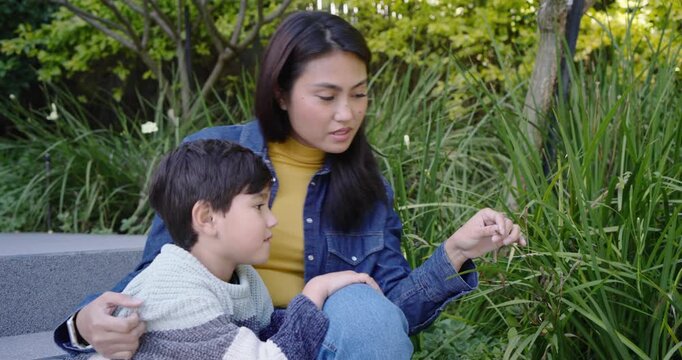 Diverse mother and son sitting on step in park, spotting tiny leaf, inspecting and learning