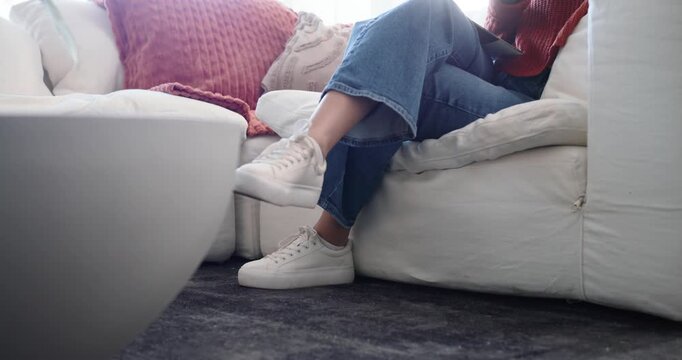 Seated woman is holding smartphone and shifting feet to avoid robot vacuum entering to clean carpet