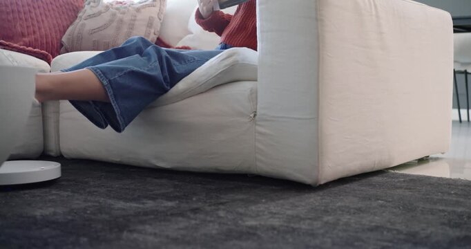 Woman sitting on right sofa reading paperback, adjusting legs as robot vacuum passes, copy space