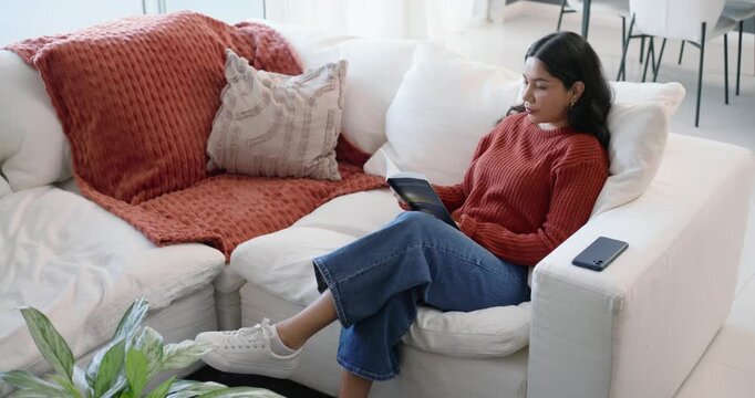 Woman sitting on sofa in orange sweater, flipping booklet while relaxing with feet on table, phone