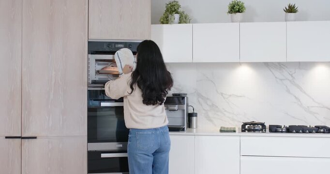 Woman reaching into open oven, removing hot baking tray with padded mitts to place on countertop