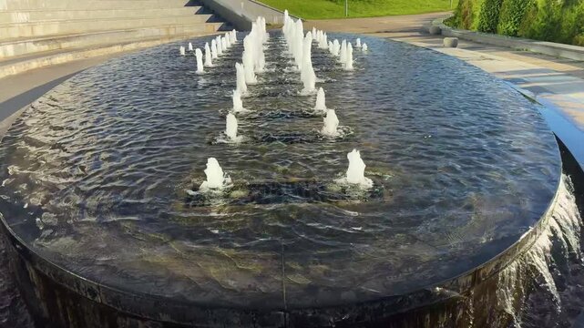 White water jets spraying from a large oval fountain beside outdoor steps, urban fountain, public space, flowing water, landscape architecture