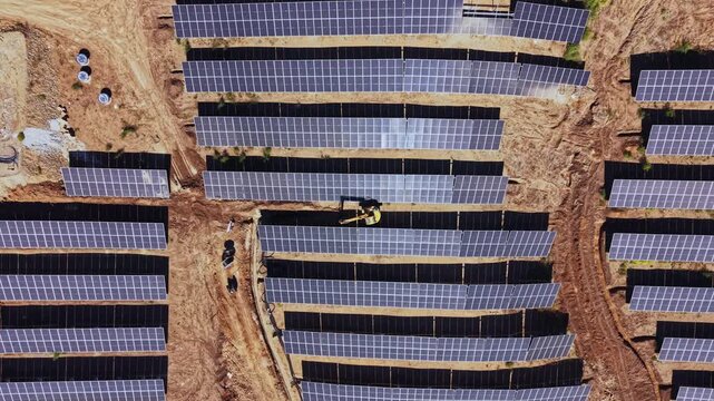 Solar panels line a ground-mounted energy site, where a worker and construction machine are positioned between rows on a dirt field during daytime.