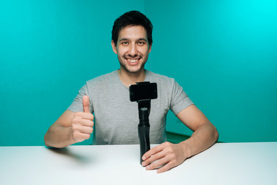 Smiling young man recording vlog with smartphone on gimbal, giving thumbs up to camera creating digital media content against turquoise studio background.