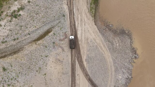 Top-down aerial tracking shot of a loaded dump truck driving on a gravel road beside a muddy river