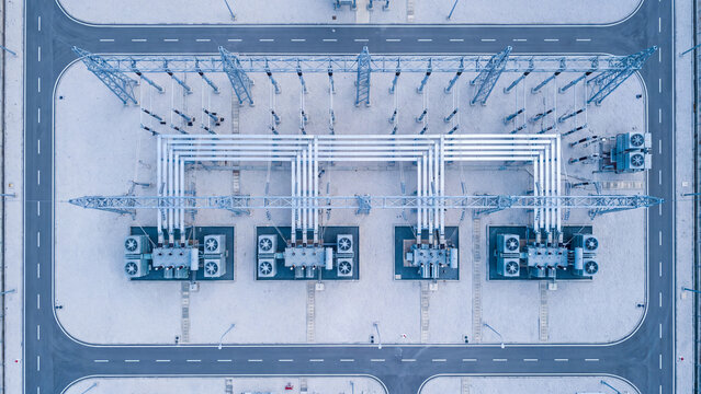Top down drone view of electrical substation with four power transformers, cooling fans and busbars surrounded by roads, high voltage grid infrastructure in cool blue tone