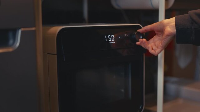 Close-up of woman's hand turning knob on portable electric oven, setting preheat mode, adjusting temperature, and pressing start button. Preparing to roast or bake food in a home kitchen.