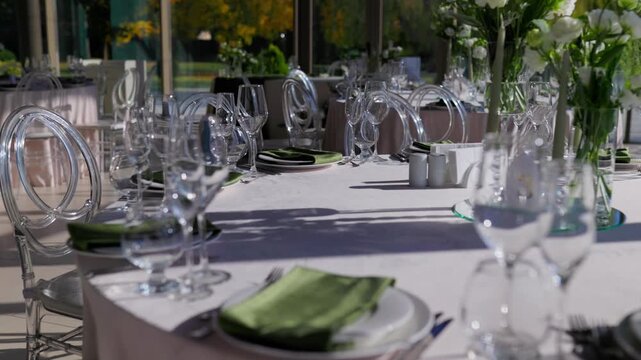 Ghost chairs around sunlit banquet table with white flowers. Modern wedding venue interior decor