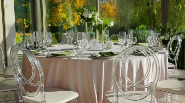 Reception table number two with floral vases and ghost chairs. Autumn park view through windows