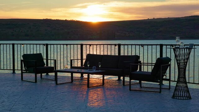 Sunset terrace lounge with cocktail glasses by lake railing. Golden sun reflection on water waves