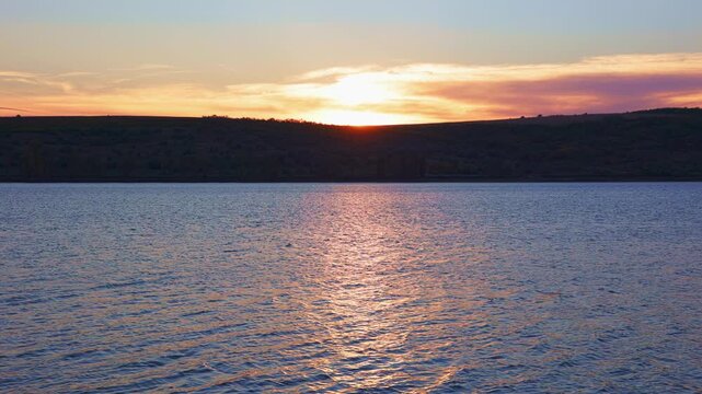 Lake sunset with golden sunbeams reflecting on rippling water. Scenic twilight over dark hills