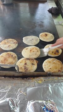 Close-up shot of a cook using a metal spatula to turn over authentic handmade pupusas filled with melted cheese and beans on a hot griddle plate. Features sizzling melted cheese, toasted corn dough, a