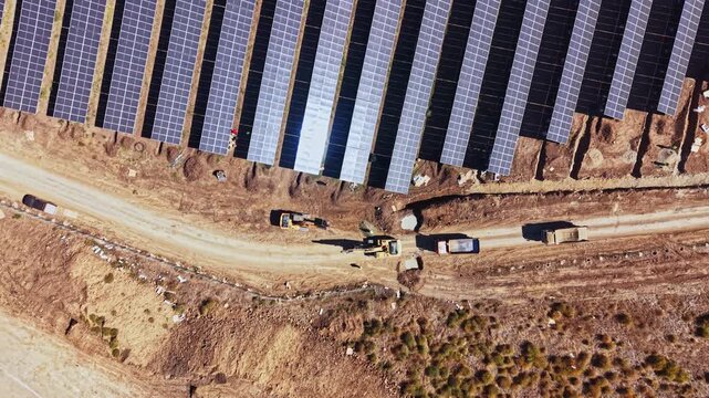 Solar panels cover a large section of land as trucks travel along a dirt road at a solar farm, seen from above during daylight.